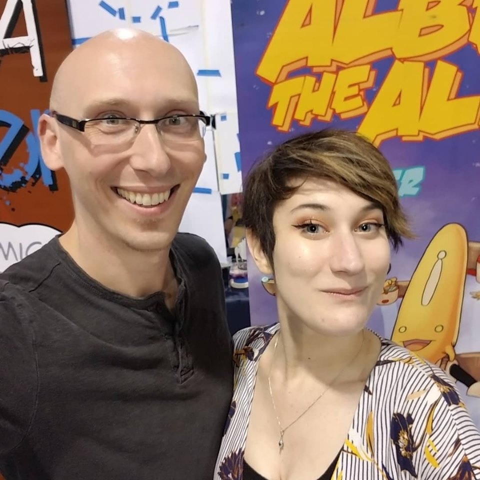 Comic writer and convention guest Trevor Mueller and volunteer Nikita Rauba at Anime Midwest 2019 albert the alien banner